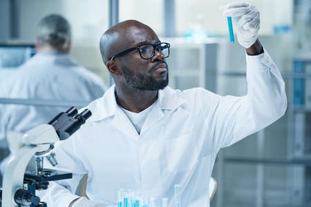 African scientist in eyeglasses and lab coat looking at flask with blue liquid in his hand during researching with microscope in laboratoryの写真素材
