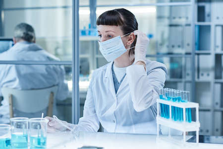 Laboratory technician in mask sitting at table in laboratory and working with samples in test tubes