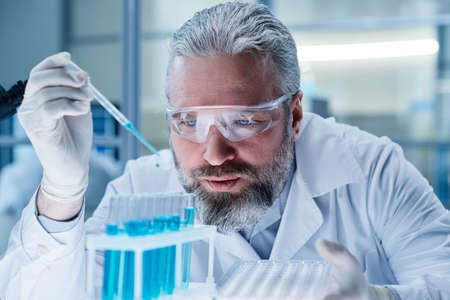Professional scientist in protective glasses adding blue liquid in tubes with pipette during medical...