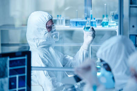 Laboratory technician in protective suit examining the blue liquid in flask while working with...