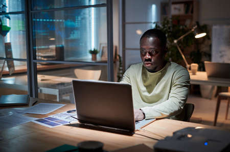 African businessman using laptop to prepare with his report sitting at table at board room and working till late eveningの写真素材