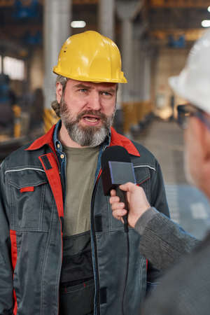 Mature male worker in hardhat and uniform talking in microphone while giving interview about machine factory to reporterの写真素材