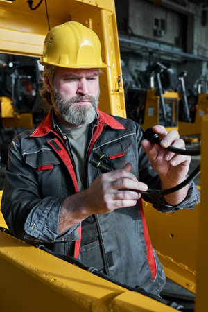 Mature bearded electrician examining the wires and connecting the equipment during his work in workshopの写真素材