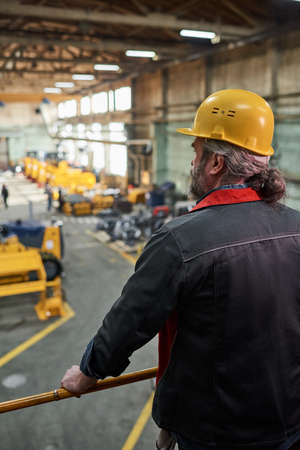 Rear view of mature foreman in work helmet standing on balcony and watching for the production...