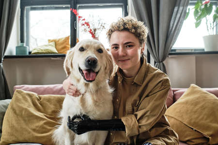 Portrait of young woman with prosthetic arm embracing her lovely golden retriever and looking at...
