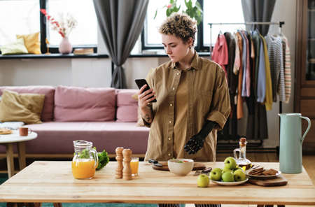 Young woman with prosthetic arm using her mobile phone to learn to prepare the food at table in room