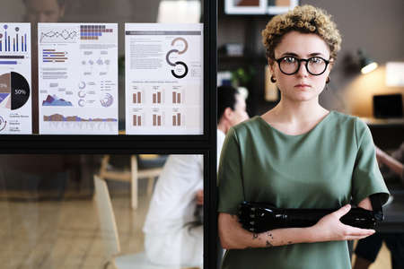 Portrait of young businesswoman with with prosthetic arm leaning on glass wall with financial...