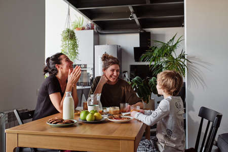 Happy lesbian couple laughing during breakfast at table while their little son talking funny jokesの写真素材