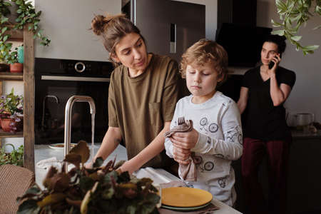 Little son wiping dishes after his mother washing them during housework in kitchen