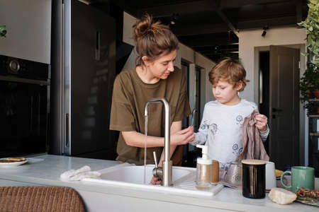 Young mother standing at sink in kitchen and washing dishes while her son helping her