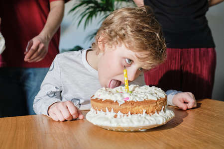 Cute little boy with blond hair tasting cream on homemade birthday cake with candle at table