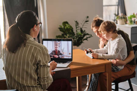 Rar view of businesswoman having online meeting with businessman on laptop, she working at home with her family in backgroundの写真素材