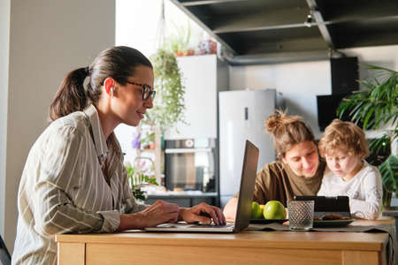 Young woman in headphones working on laptop at table with her family playing in backgroundの写真素材