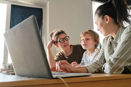 Two lesbian mothers talking to their son while sitting at table in front of laptop during online lessonの写真素材
