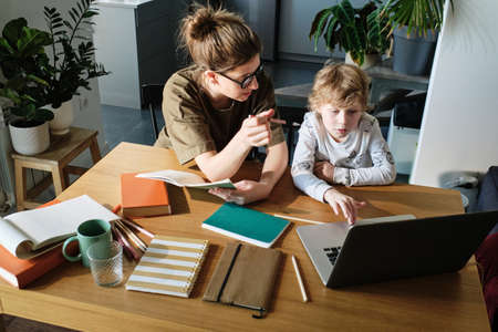 Little boy using laptop and doing online work together with tutor while they sitting at table at...
