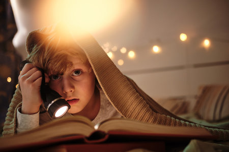 Little boy lying under blanket on bed and looking at camera while reading a book with flashlightの写真素材