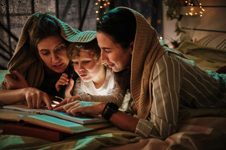 Homosexual mothers lying on bed under blanket with their son and reading a book together using...