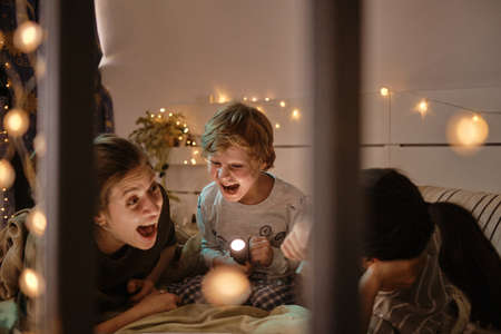 Excited little boy with flashlight laughing together with his homosexual mothers while they lying on bed in bedroomの写真素材