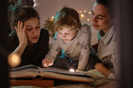 Little boy reading a tale with his homosexual mothers together before sleep while lying on bed