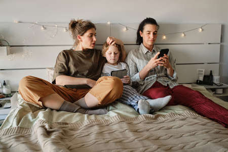 Lesbian family resting on bed in bedroom with their little son and using gadgetsの写真素材