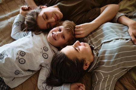 High angle view of little boy smiling at camera while lying on bed with his homosexual mothersの写真素材