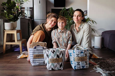 Portrait of homosexual mothers with little son looking at camera sitting on the floor with bags with sorted garbage for recyclingの写真素材