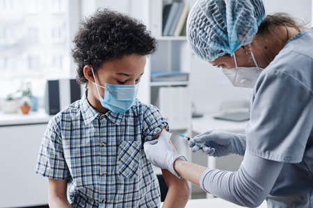 Pediatrician in mask giving vaccination in arm of little child during his visit at hospitalの写真素材