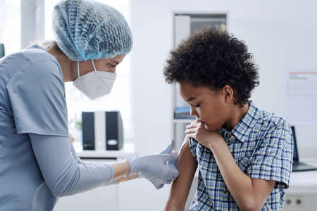 Female nurse in mask applying bandage on shoulder of little boy after vaccination at hospitalの写真素材