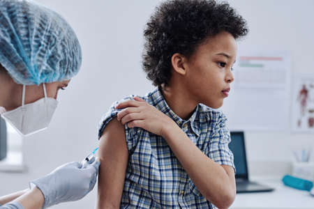 African little boy looking away while doctor injecting his shoulder with vaccine during visit at hospitalの写真素材