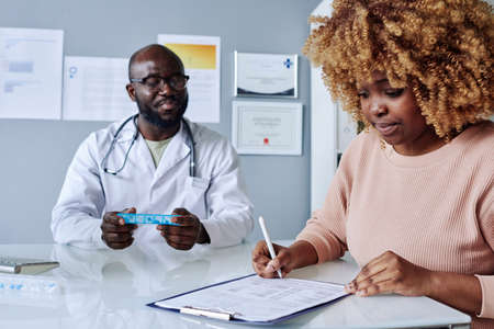 African young woman with curly hair signing insurance before treatment while sitting at doctors...