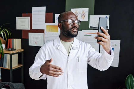 African male doctor in white coat using mobile phone for online consultation during work at officeの写真素材