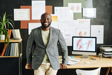 Portrait of successful medical specialist smiling at camera while standing near his workplace with...