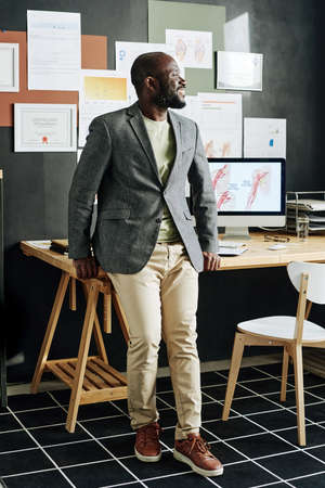 Successful African physician smiling and looking away while standing at his office with computer and...