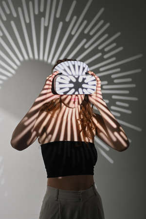 Girl in goggles playing virtual reality game standing against the grey background with bright pattern light on herの写真素材