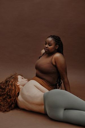 African plump woman sitting with blank expression with other young woman lying on her knees isolated on brown backgroundの写真素材