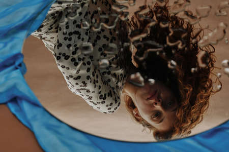 Young red haired woman looking at mirror with sad expression through drops on itの写真素材
