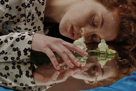 Young woman lying with her eyes closed and touching the flower lying on wet mirrorの写真素材