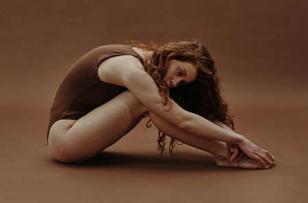 Young pretty woman with red hair sitting in swimsuit and stretching against the brown backgroundの写真素材