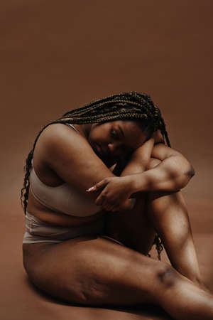 Portrait of African plump woman in underwear sitting against the brown background and looking very sadの写真素材