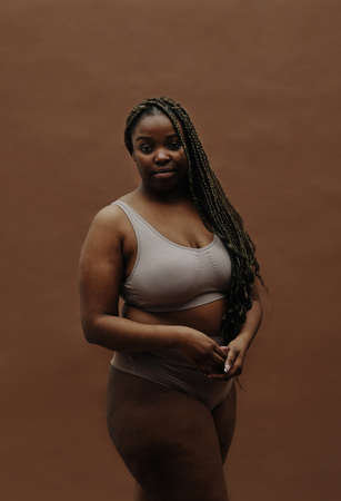 Portrait of African young woman with long braids having plump figure standing in underwear against...