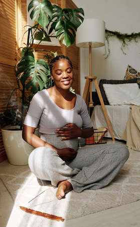 African happy woman sitting on floor in bedroom and touching her pregnant belly, she enjoying her...