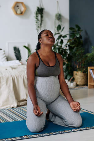 African pregnant woman sitting on exercise mat and deeply breathing during meditation at homeの写真素材