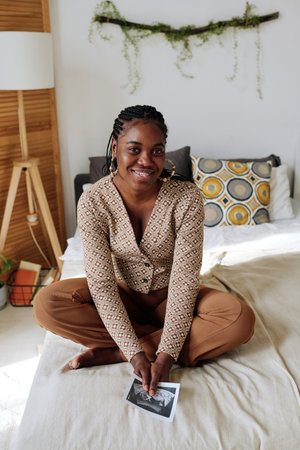 Portrait of African young woman sitting on bed and smiling at camera, she learning about her pregnancyの写真素材