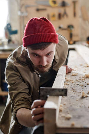 Vertical shot of serious young male carpenter using steel square when working with wood plank in woodshopの写真素材