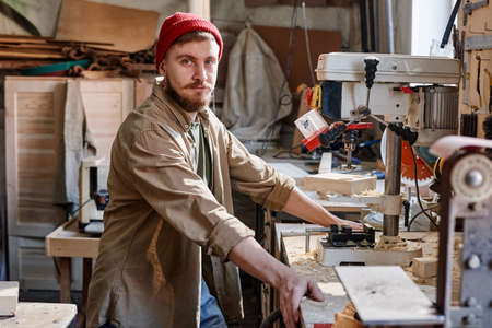 Portrait of young Caucasian male carpenter with beard on face working with drill press looking at...