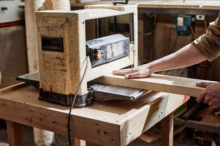Unrecognizable male carpenter working using thickness planer machine to trim wood plank in...