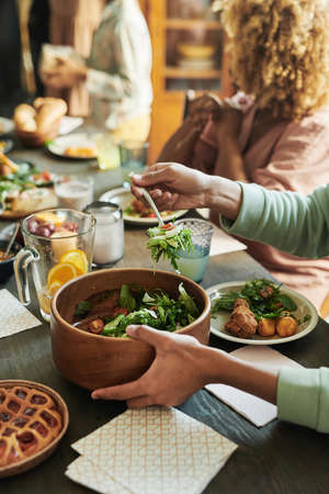 Close-up of boy putting fresh vegetable salad from bowl on his plate during dinner at table with his familyの写真素材