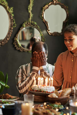 African grandfather sitting at table with closed eyes while his granddaughter congratulating him with birthday with cakeの写真素材