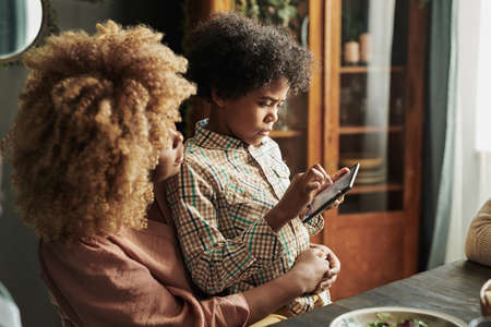 African little son using mobile phone while sitting on knees of his mother while they having dinner at tableの写真素材