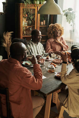 Big African family sitting at table and talking for cup of tea and cake in dining room